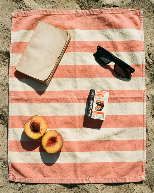 A box of Energy Strips on a striped beach towel in the sand, with sunglasses, a book and a halved peach
